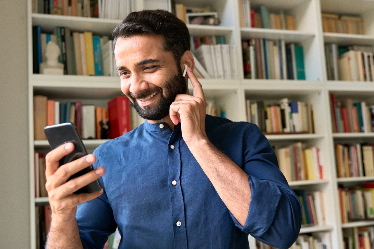 wo colleagues in a bright room smiling and looking at a laptop. The woman has curly short hair and a brown shirt, while the man has a beard and a light shirt.